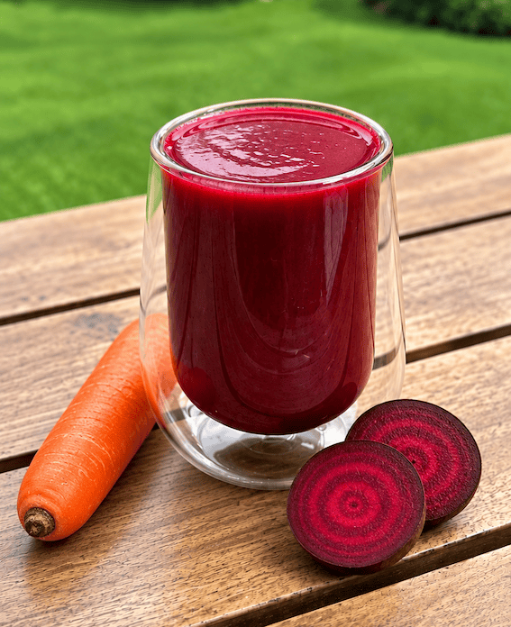 Un vaso de jugo rojo brillante junto a una zanahoria y rodajas de remolacha sobre una mesa de madera.