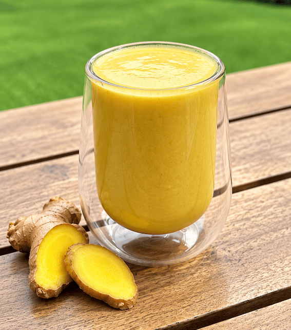 Vaso transparente con un smoothie amarillo en una mesa de madera, acompañado de rodajas de jengibre fresco.